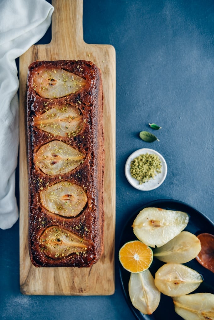 Pear bread on a wooden board accompanied by caramelized pears and a wedge of orange in an iron pan on the side.