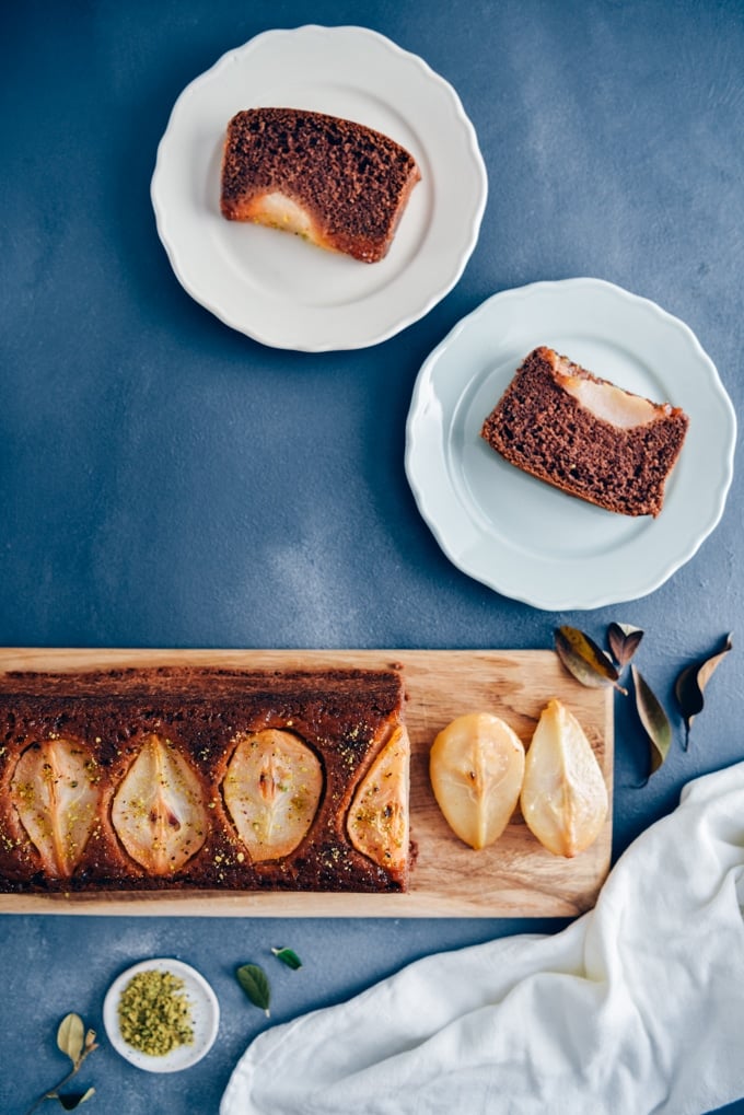 Pear loaf cake sliced on a wooden board accompanied by two plates, a small bowl of pistachio.