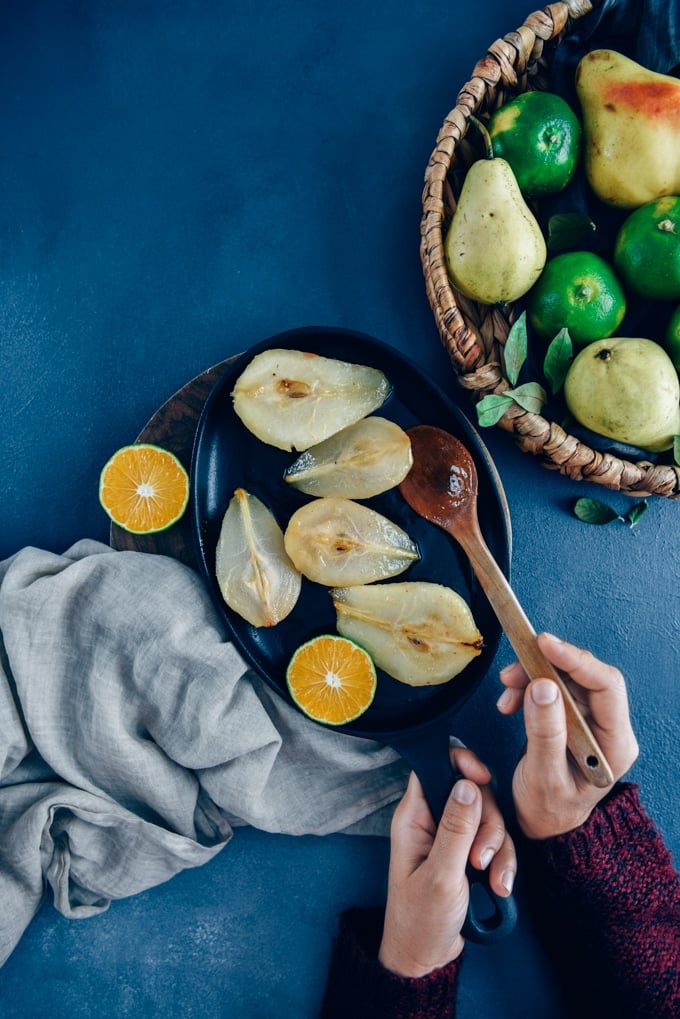Hands holding a wooden spoon and an iron pan with caramelized pears and a wedge o orange inside. Fresh pears and oranges in a basket on the side.