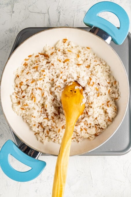 Browned orzo and rice are in a white pan with a wooden spoon in it.