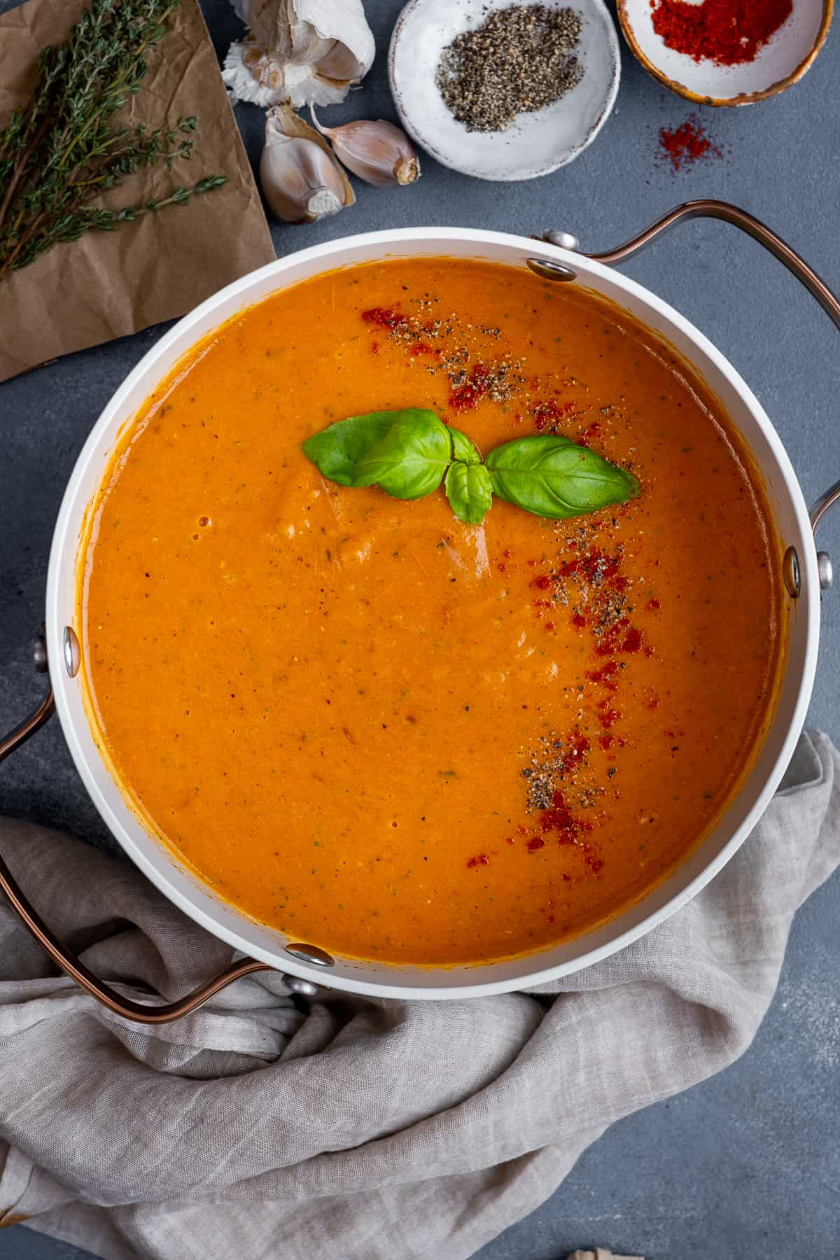 Roasted tomato soup garnished with spices and fresh basil leaves in a pot, black pepper, paprika, garlic and thyme on the side.