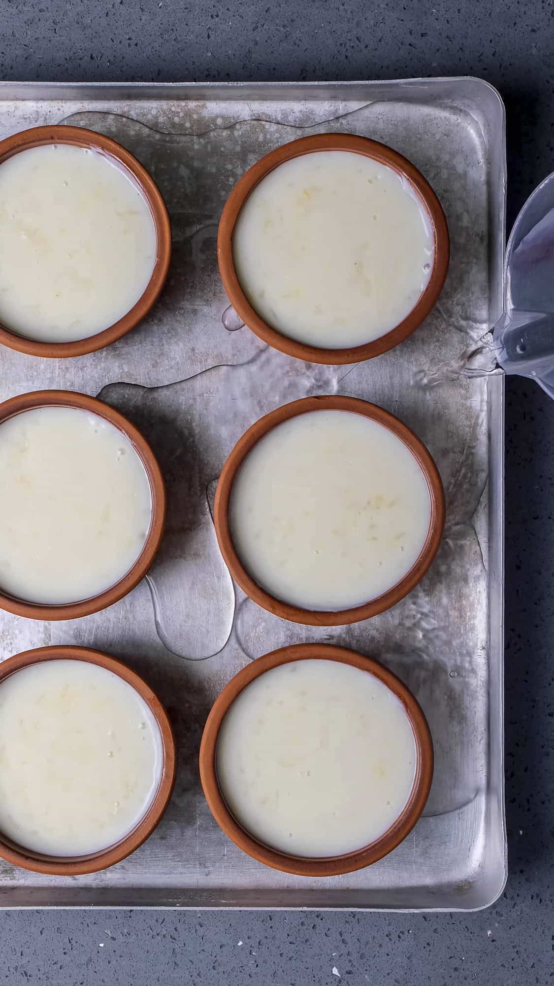 Pouring water from a jug into an oven tray with rice pudding bowls in it.