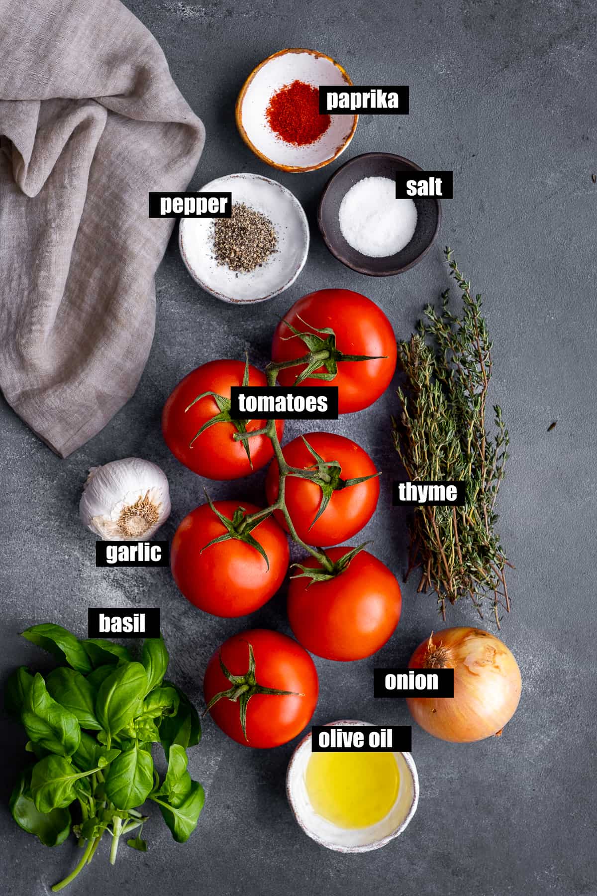 Tomatoes, onion, garlic, oil, basil, thyme, paprika, salt and pepper on a dark grey backdrop.