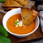 Roasted tomato soup topped with feta cheese and basil leaves in a white bowl, grilled cheese sandwich dipped into it and more grilled cheese sandwiches behind it.