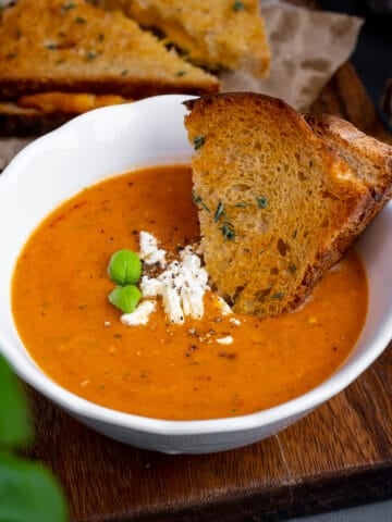 Roasted tomato soup topped with feta cheese and basil leaves in a white bowl, grilled cheese sandwich dipped into it and more grilled cheese sandwiches behind it.