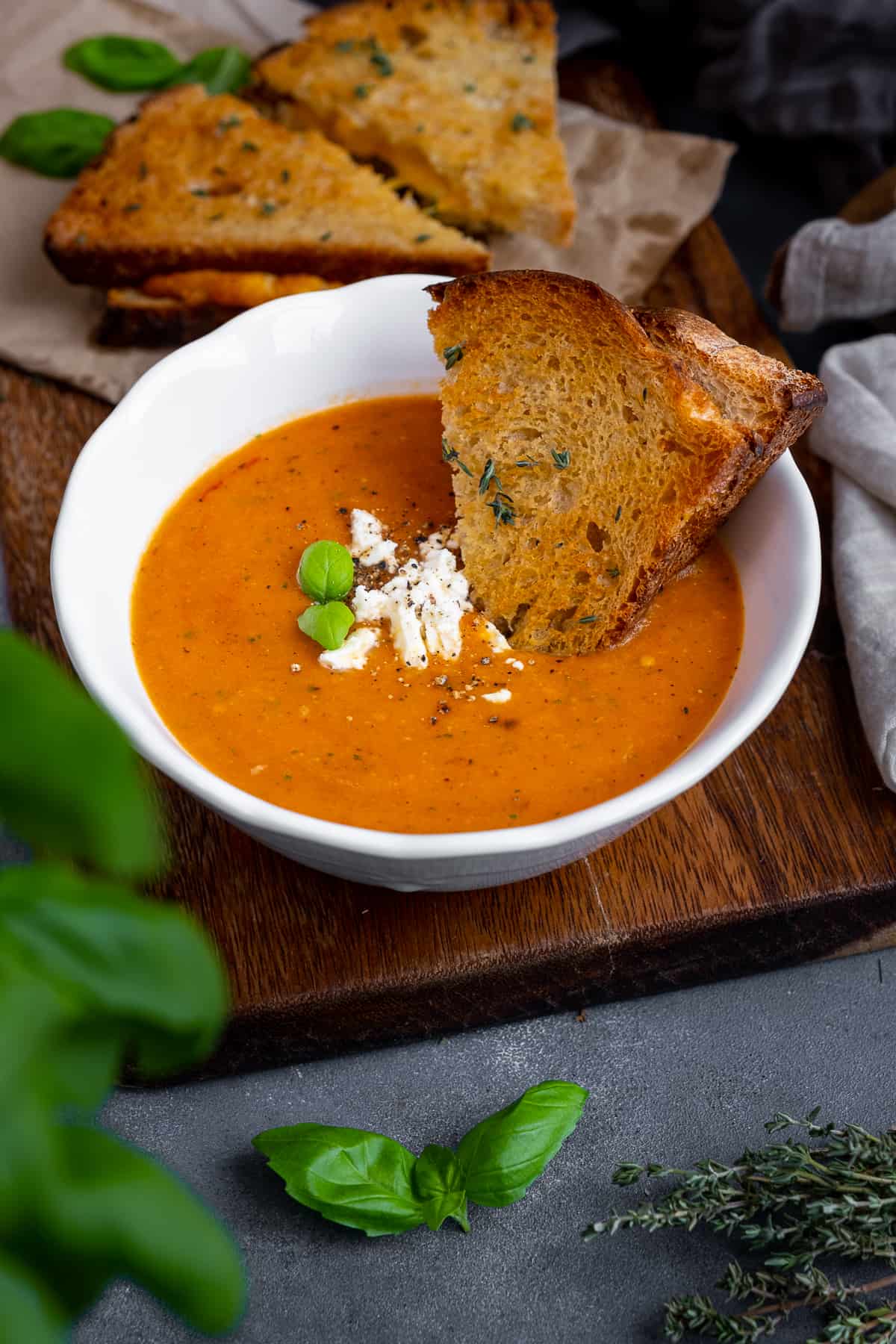 Roasted tomato soup topped with feta cheese and basil leaves in a white bowl, grilled cheese sandwich dipped into it and more grilled cheese sandwiches behind it.