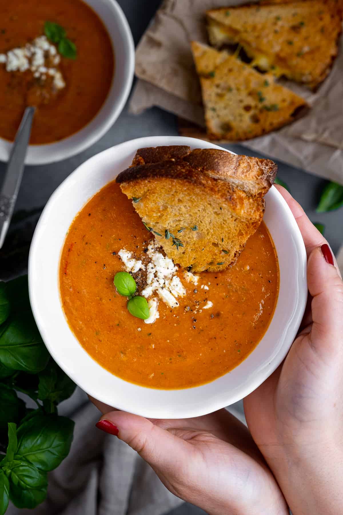 Woman hands holding a bowl of tomato soup topped with feta cheese and basil leaves and a grilled cheese sandwich dipped into it.