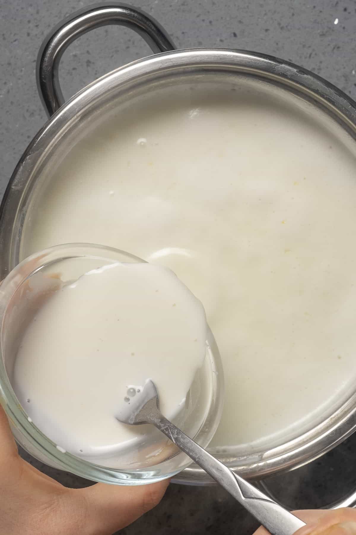 Hands pouring a mixture of milk and cornstarch into simmering milk.