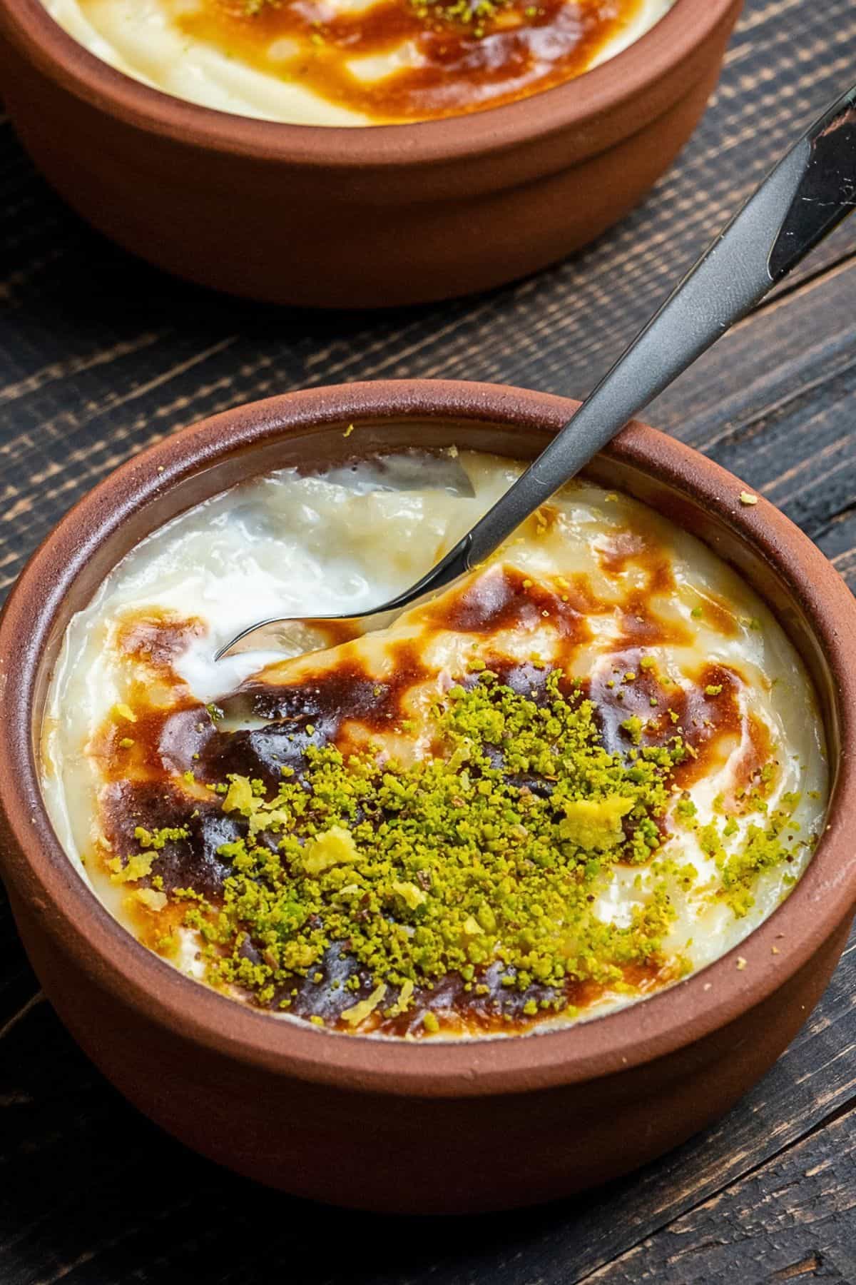 Turkish baked rice pudding (fırın sütlaç) topped with ground pistachios in a traditional clay bowl with a spoon scooping from it.