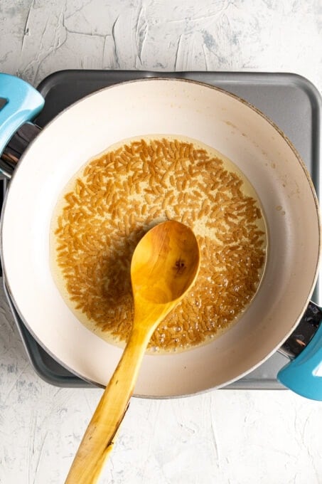 Browned orzo pasta in a white pan with olive oil and a wooden spoon in it.