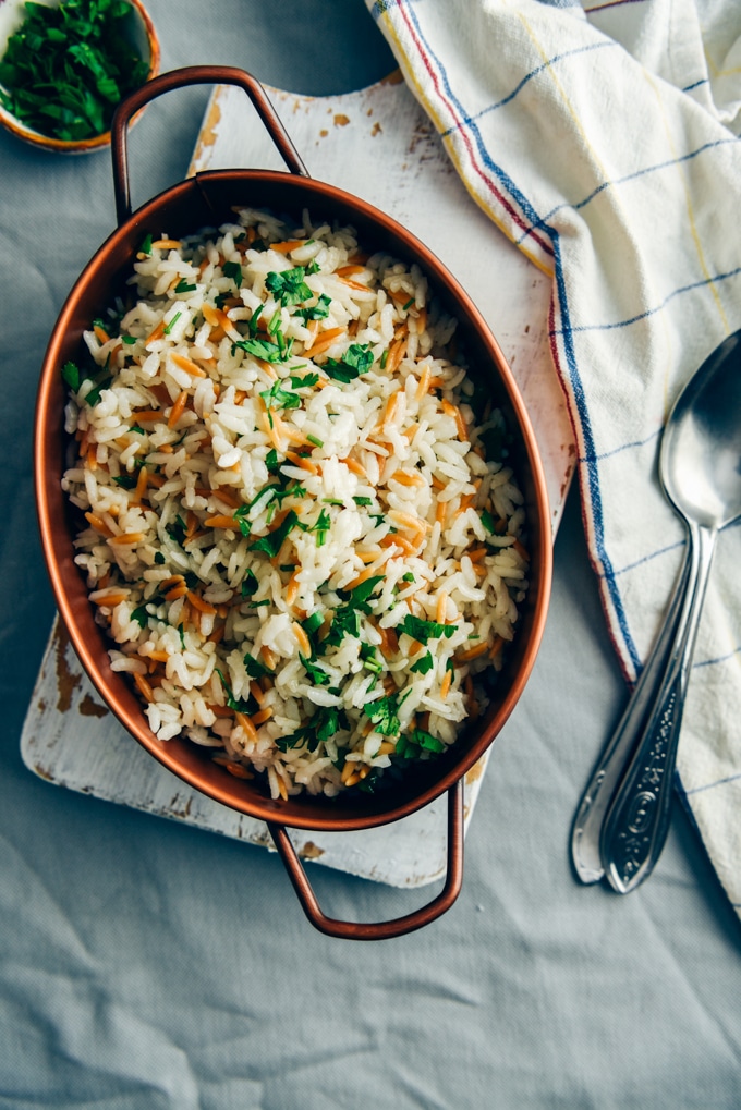 Rice pilaf with orzo served in a copper pan with two spoons on the side.