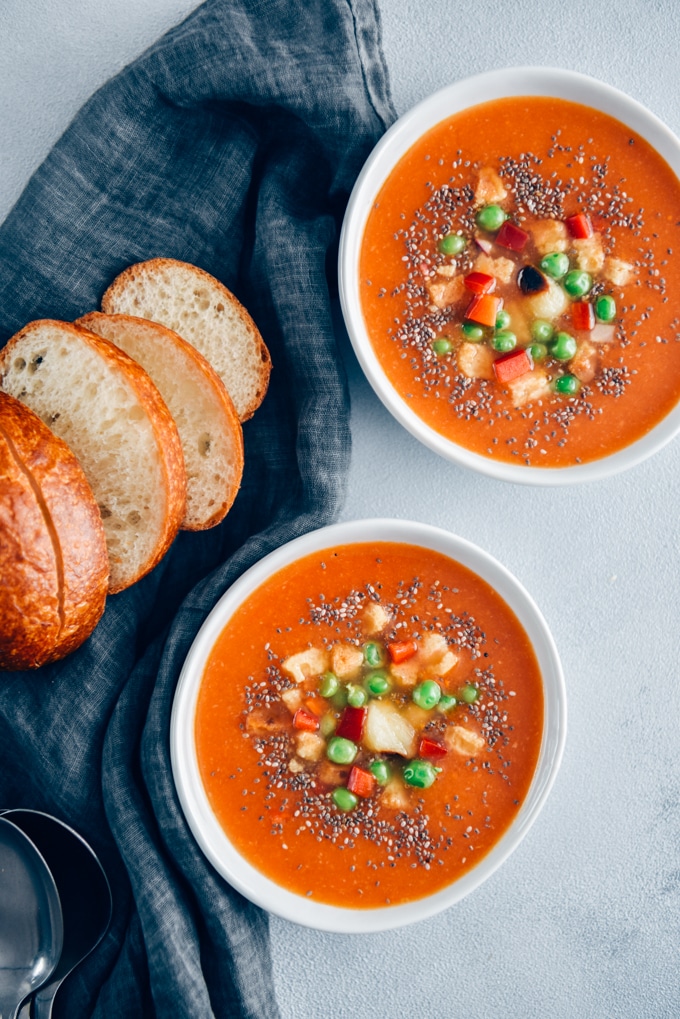 Vegan tomato soup served in two white bowls accompanied by bread rolls, roasted garlic, croutons and peas.