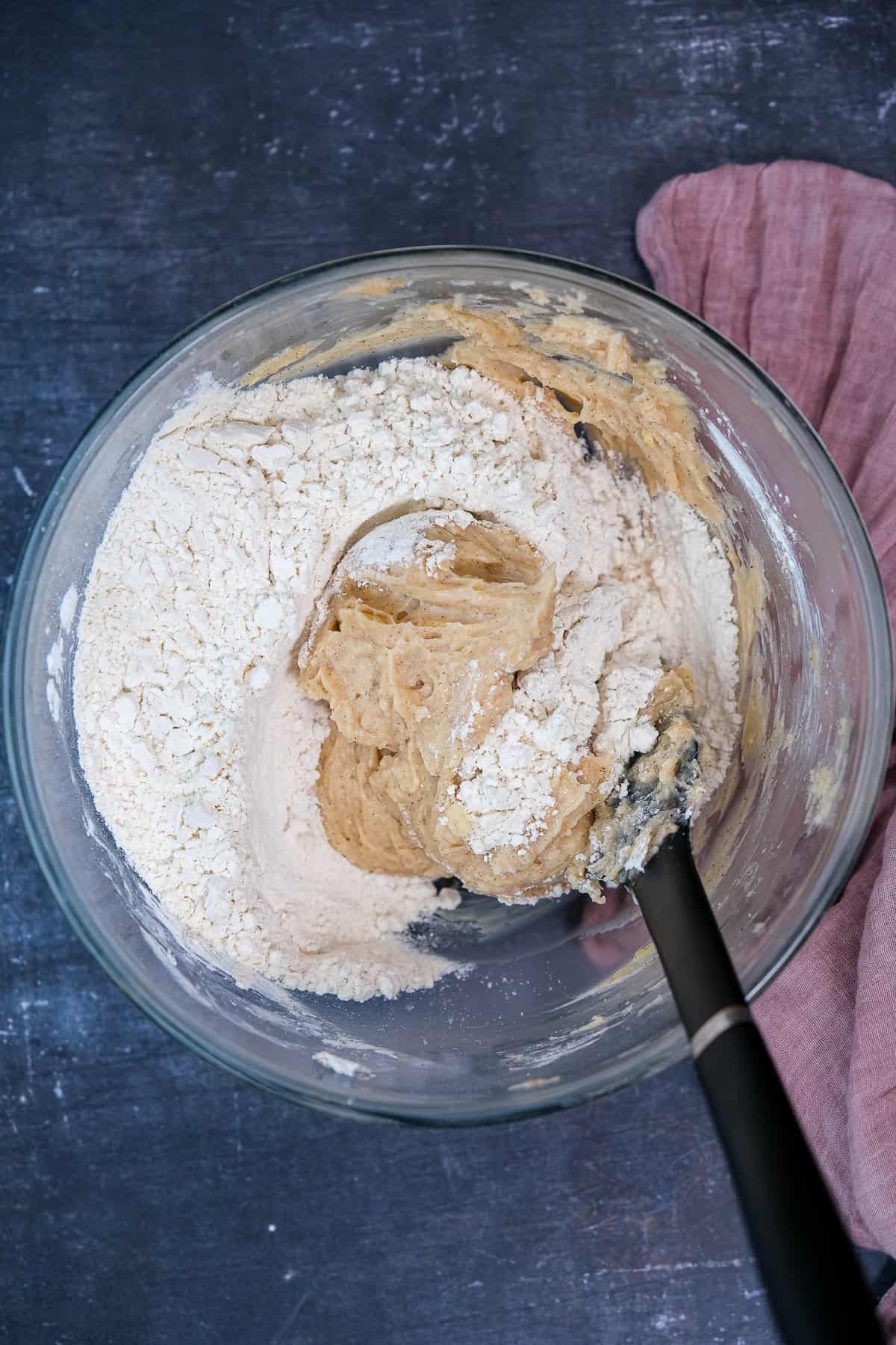 Flour is being mixed with the other ingredients in a glass mixing bowl and a black spatula inside it.
