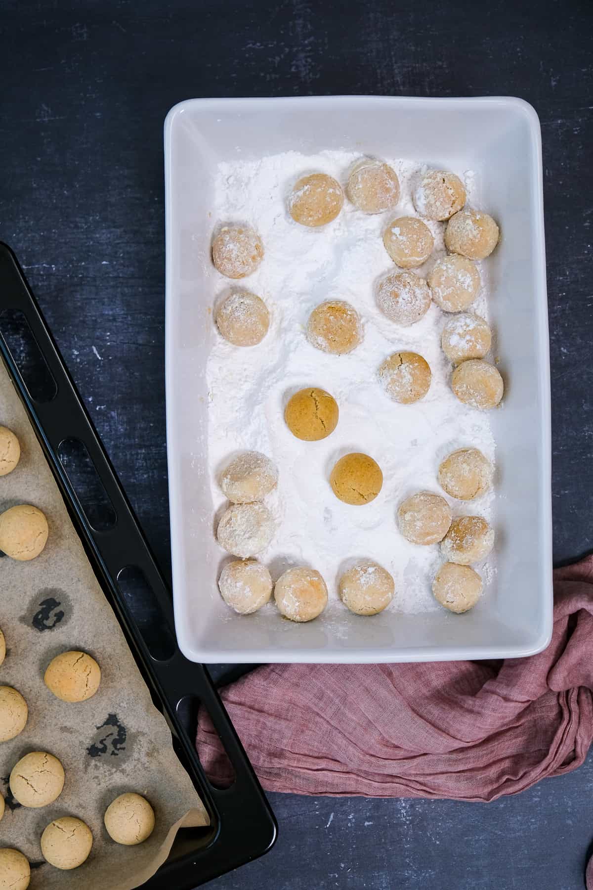 Mini cookie balls are being coated with powdered sugar in a rectangular white pan, more cookies on a baking sheet on the side.