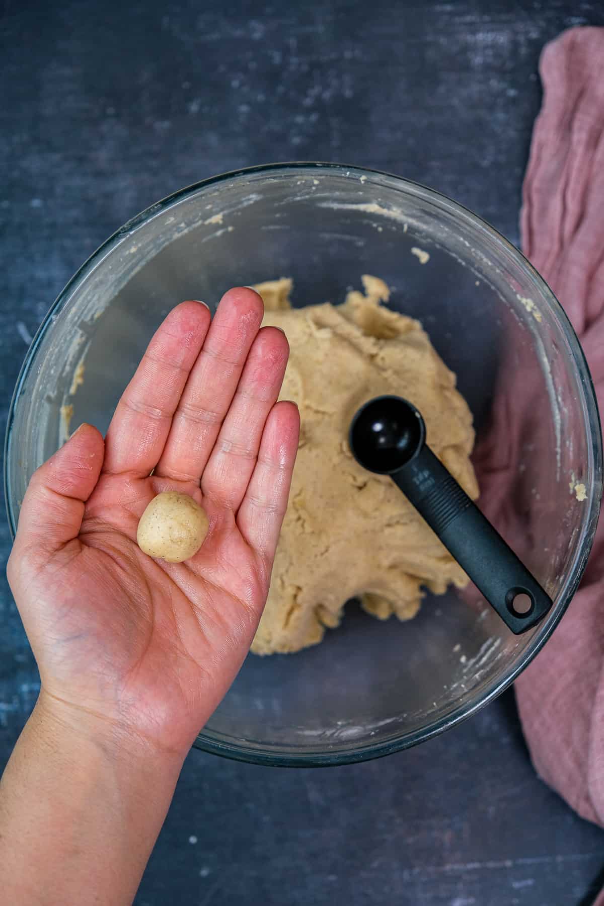 A hand holding a mini cookie dough ball and the rest of the dough and a teaspoon in a mixing bowl below it.