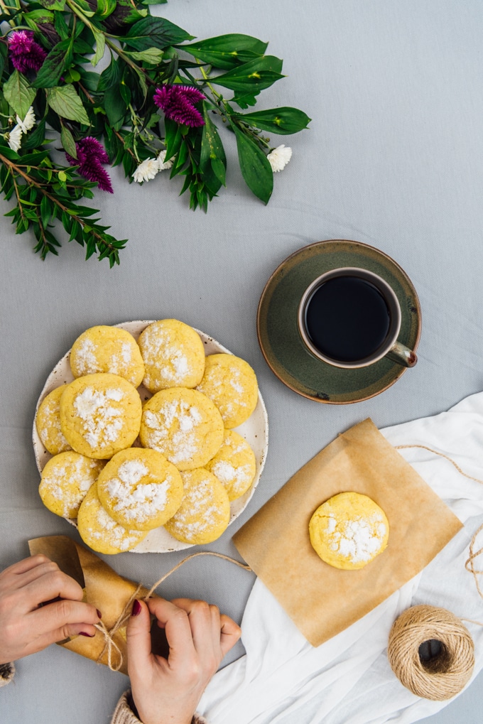 Hands wrapping lemon crinkle cookies photographed with a plate of cookies and a cup of coffee on the side