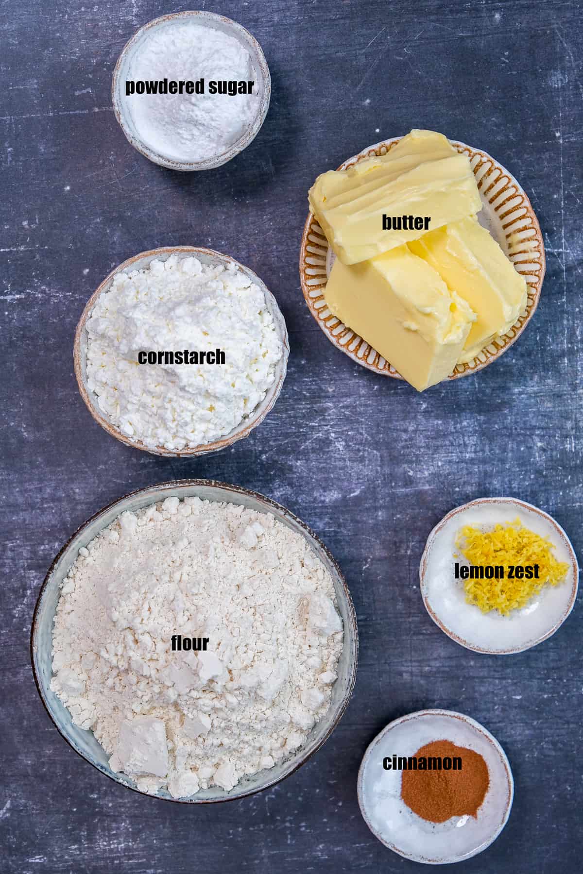 Flour, cornstarch, softened butter, lemon zest, cinnamon and powdered sugar all in separate bowls on a dark background.