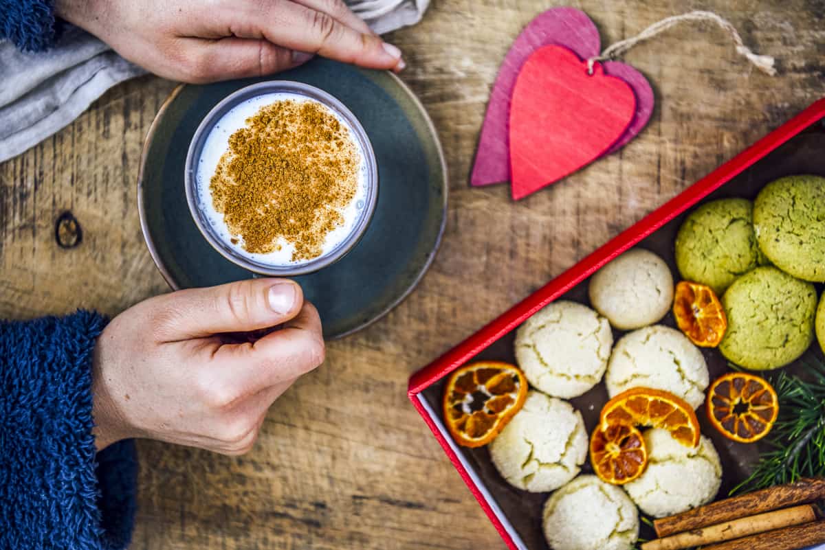 Hands holding a cup of salep garnished with cinnamon, cookies in a box on the side.
