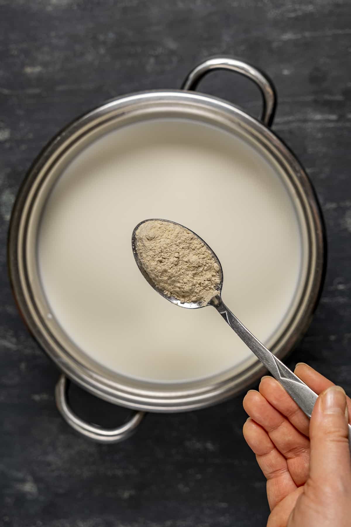 A hand holding a spoon full of salep powder over a pot of milk.