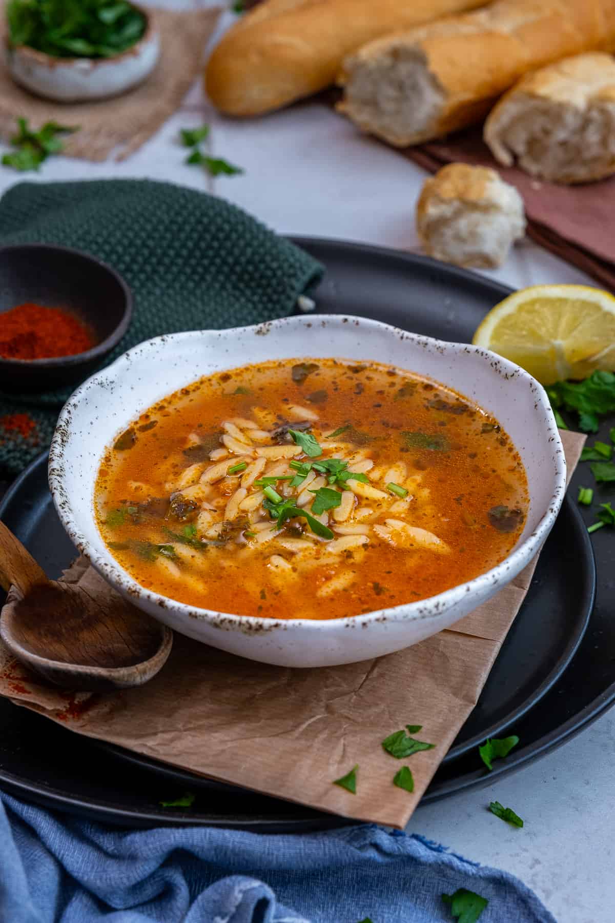 A bowl of orzo soup from front view, a wooden spoon, a lemon wedge, paprika in a mini bowl and some bread and parsley accompany.