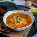 A bowl of orzo soup on a black plate, a wooden spoon, paprika in a mini bowl and a lemon wedge accompany.