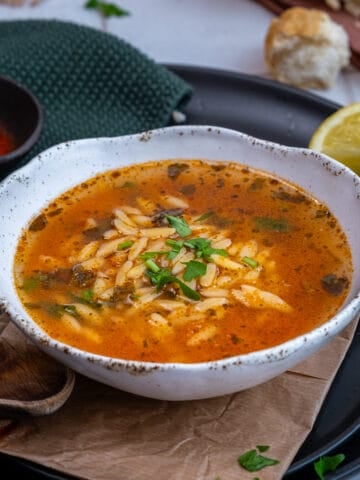 A bowl of orzo soup on a black plate, a wooden spoon, paprika in a mini bowl and a lemon wedge accompany.