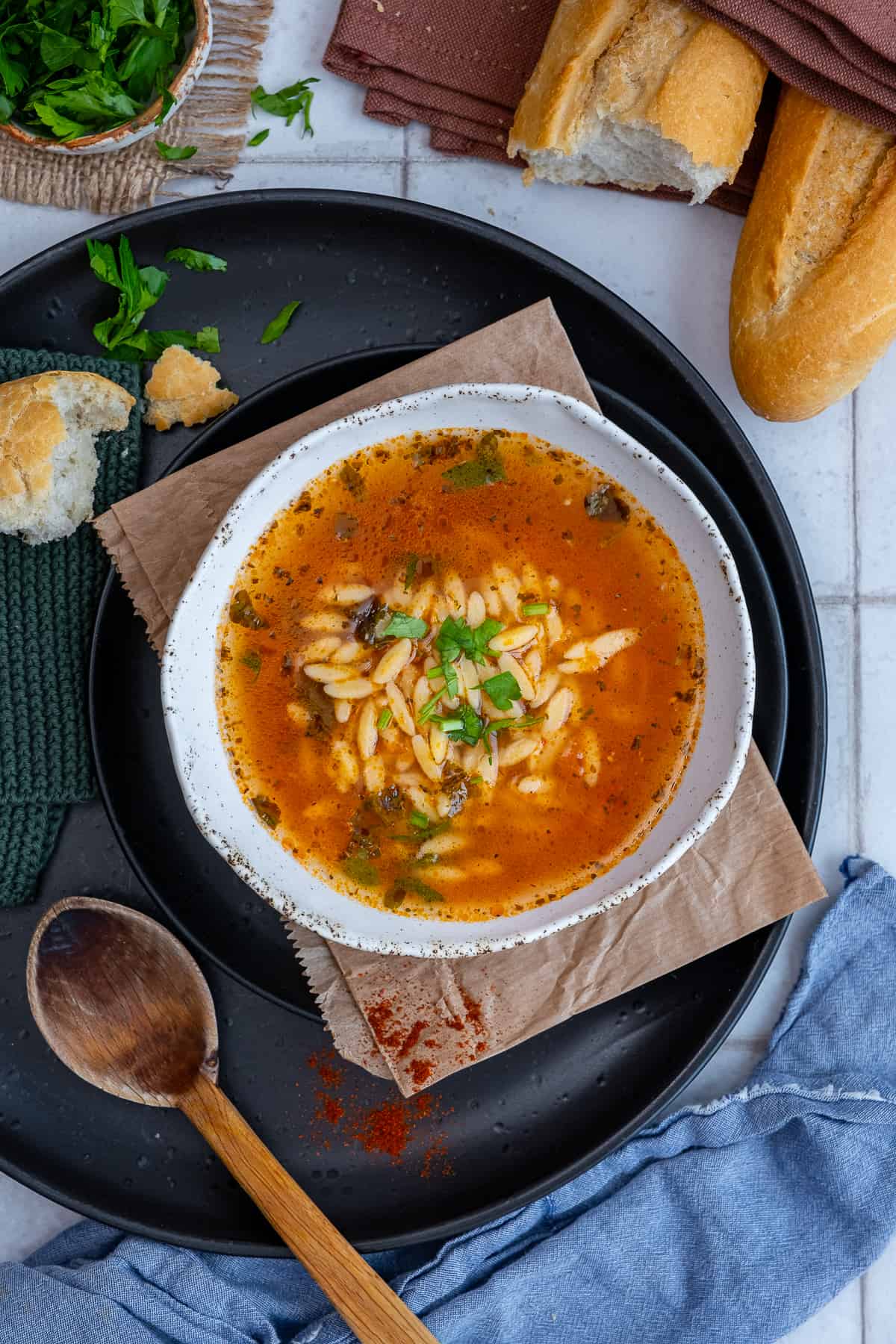 A bowl of orzo soup on a black plate, a wooden spoon on the side, some bread and parsley accompany.