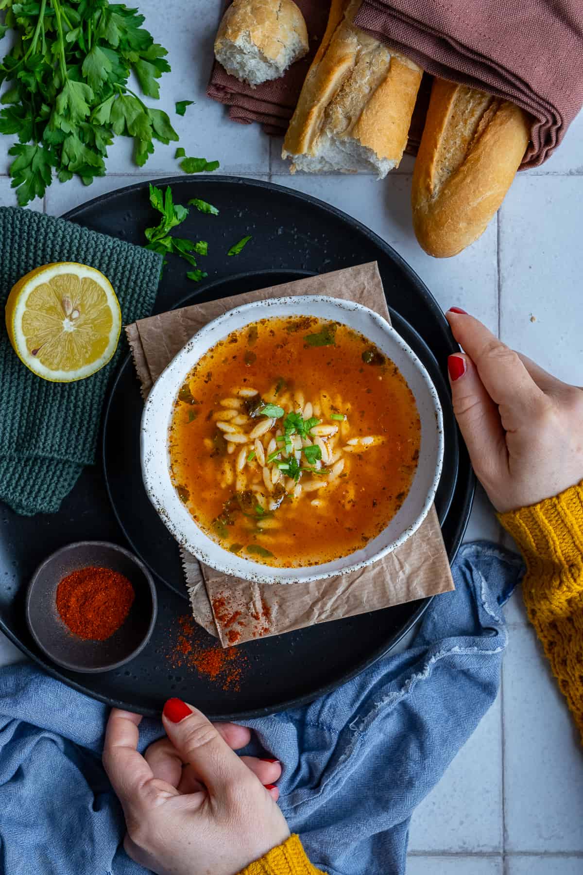 Woman hands holding a black colored plate, a bowl of orzo soup, a mini bowl of paprika and half of a lemon on it, some bread and parsley on the side.