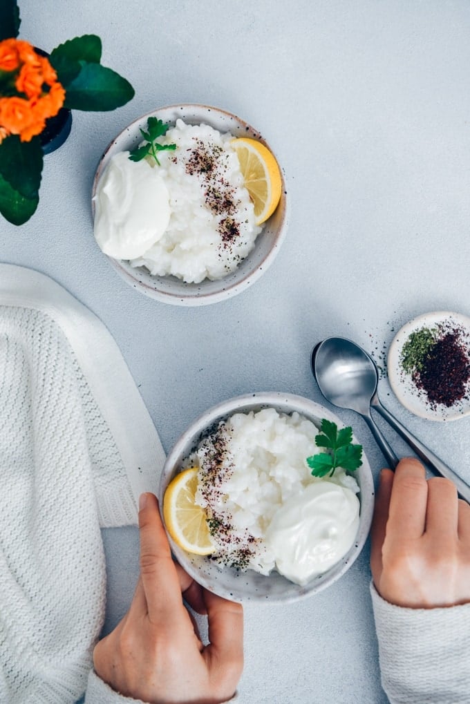 Woman eating savory rice porridge garnished with yogurt, lemon wedges, sumac and dried mint.