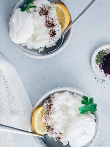 Savory rice porridge in two ceramic bowls garnished with yogurt, sumac, dried mint and lemon wedges photographed on a grey background.