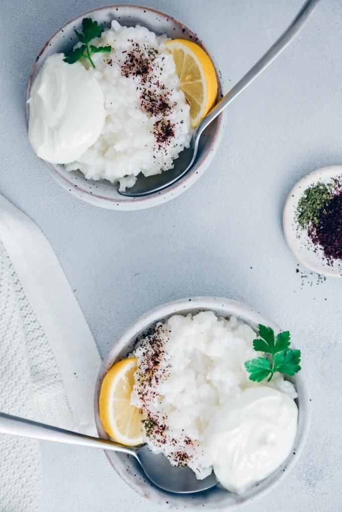 Savory rice porridge in two ceramic bowls garnished with yogurt, sumac, dried mint and lemon wedges photographed on a grey background.