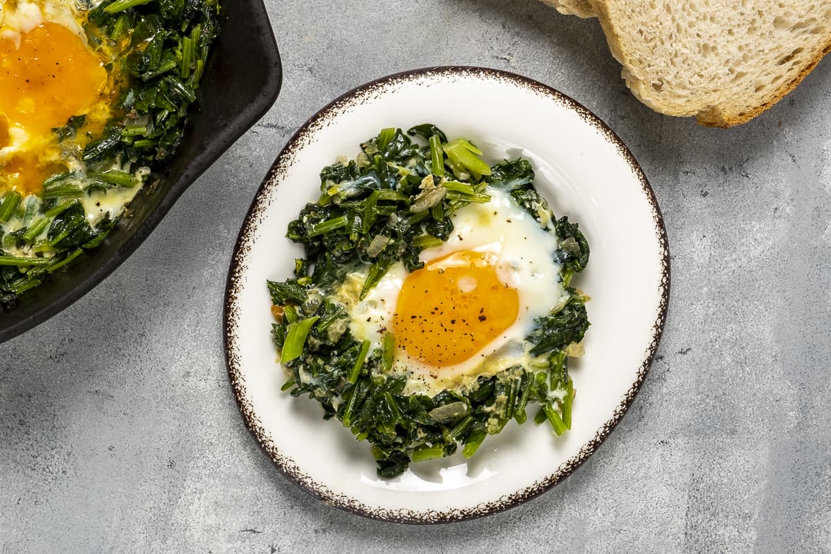 Spinach topped with an egg served on a white plate and bread slices on the side.