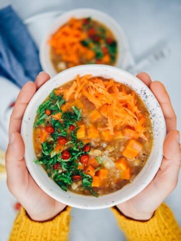 A woman with yellow cardigan holding a bowl of vegan carrot soup with rice, lentils and celeriac, garnished with mint and chili peppers.