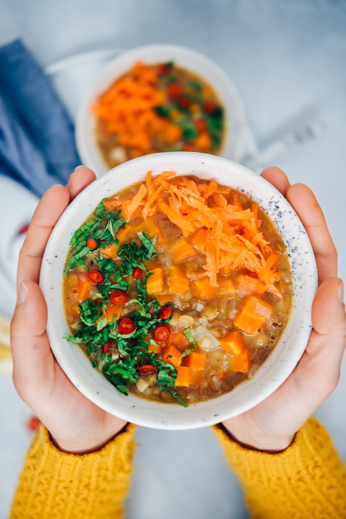 A woman with yellow cardigan holding a bowl of vegan carrot soup with rice, lentils and celeriac, garnished with mint and chili peppers.