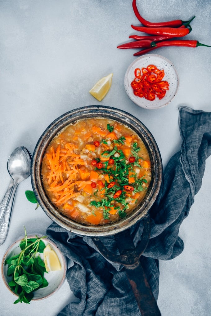 Carrot soup with lentils and rice in a vintage pan garnished with chopped fresh mint and chili peppers photographed on a light grey background from top view.