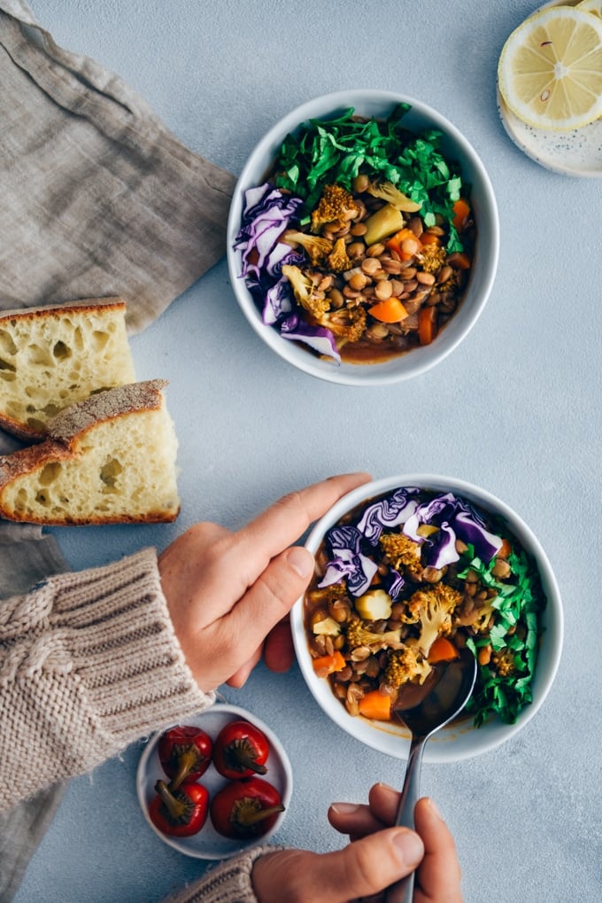 Woman eating vegan lentil stew with broccoli and carrot garnished with shredded red cabbage and chopped parsley served in white bowls. 