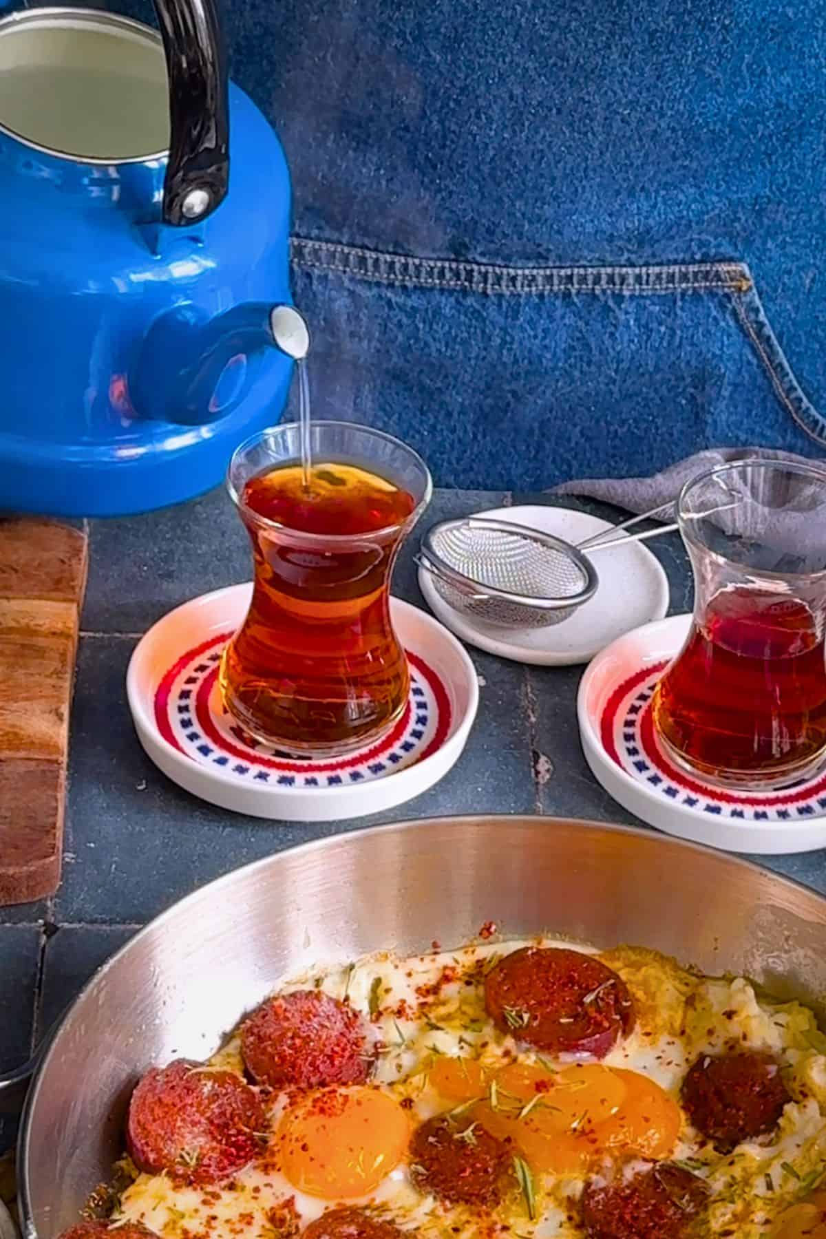 Hot water being poured from a blue Turkish teapot into a tulip-shaped glass of Turkish tea, served alongside a traditional Turkish breakfast.