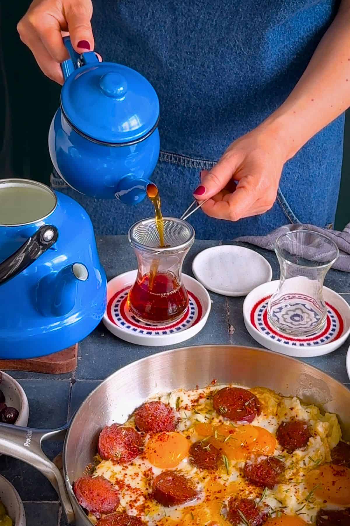 Brewed Turkish black tea being poured through a strainer into a traditional tulip-shaped tea glass, alongside a Turkish breakfast.