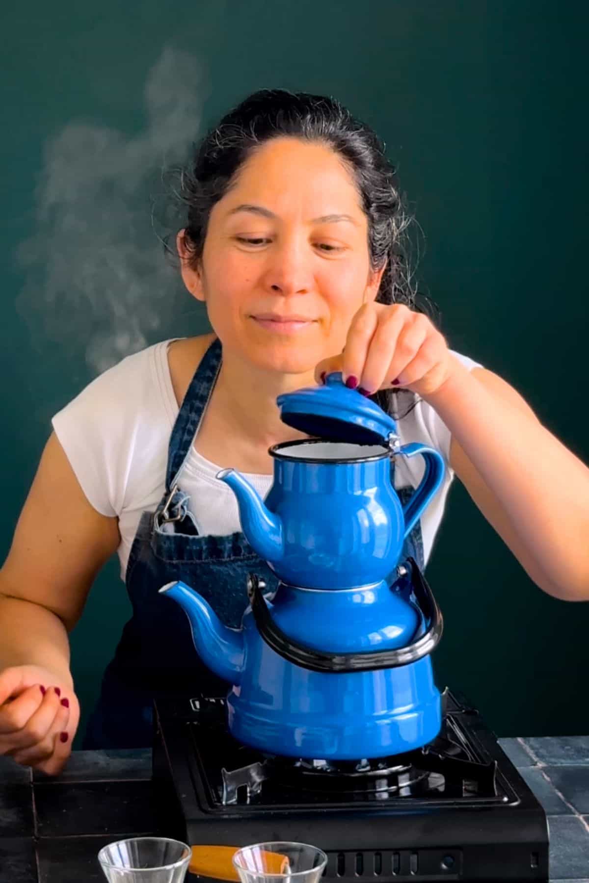Woman lifting the lid of a blue Turkish double teapot (çaydanlık) to check the brewed tea.