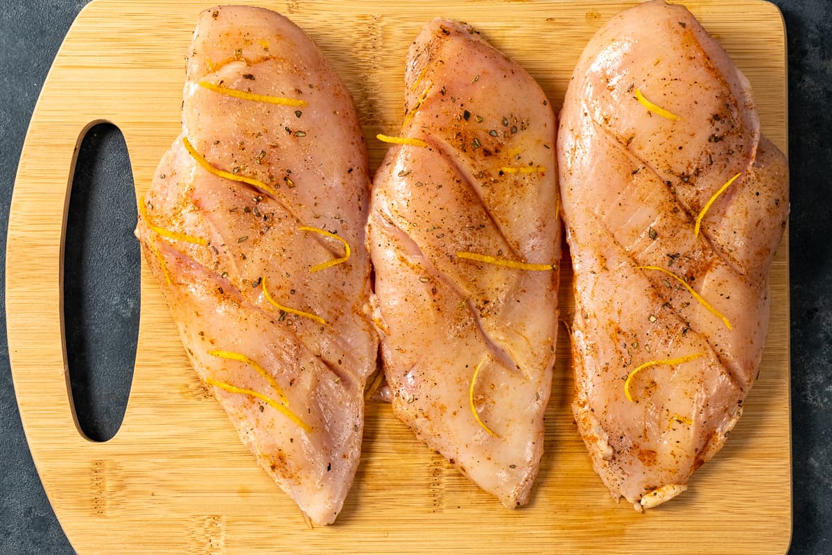 Three chicken breasts with thin slits on each and seasoned with spices and lemon zest on a wooden board.