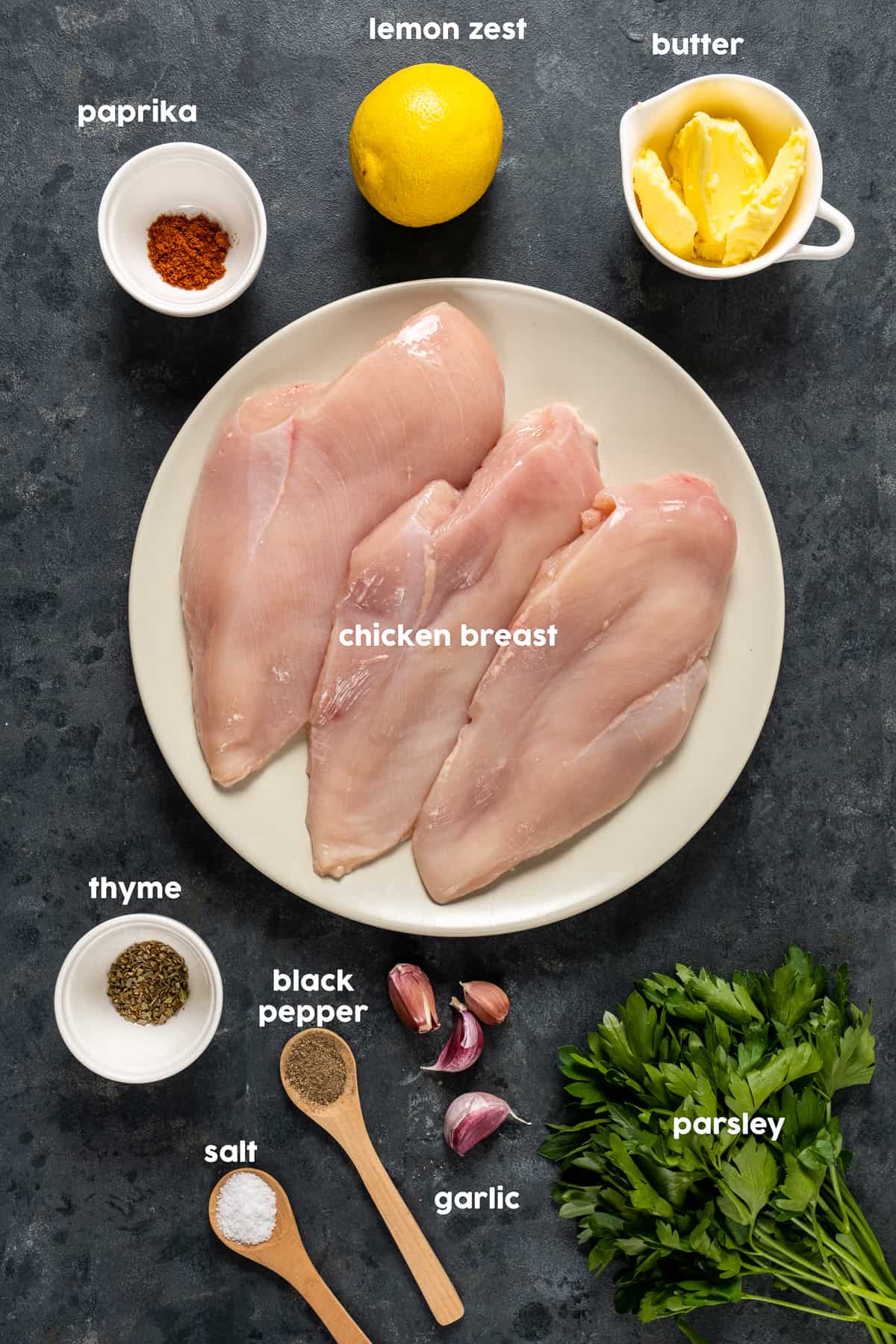 Chicken breasts on a white plate surrounded by thyme, black pepper, garlic cloves, parsley, butter, lemon and paprika on a dark background.