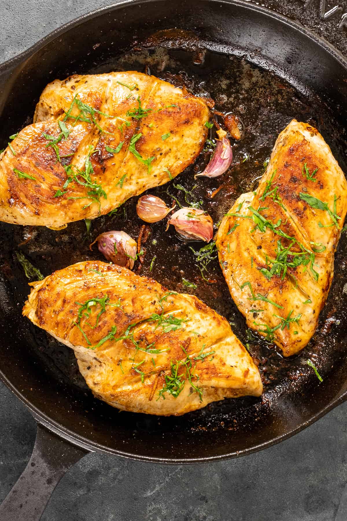 Cooked chicken breasts garnished with chopped parsley and garlic cloves in a cast iron skillet.