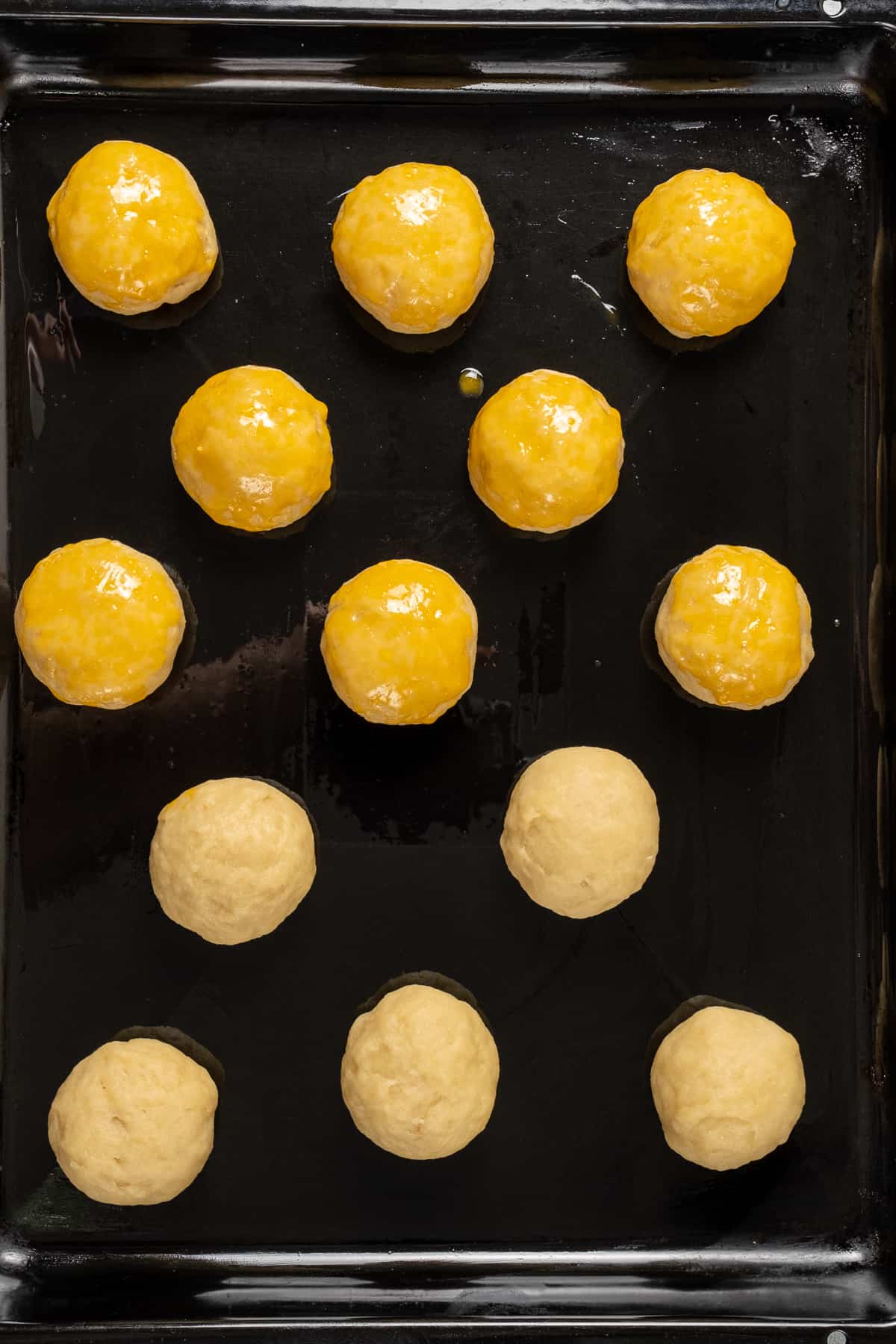 Dough balls on a greased baking sheet, half of them brushed with egg yolk.