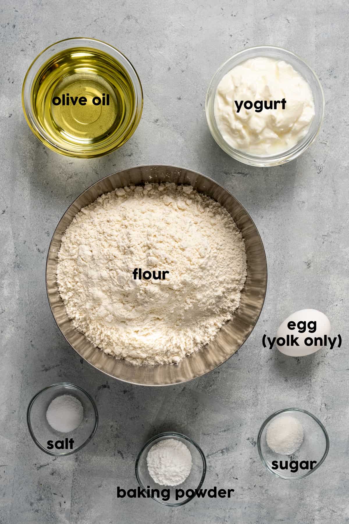 Flour, olive oil, yogurt, salt, sugar, baking powder in separate bowls on a grey background and an egg accompany.