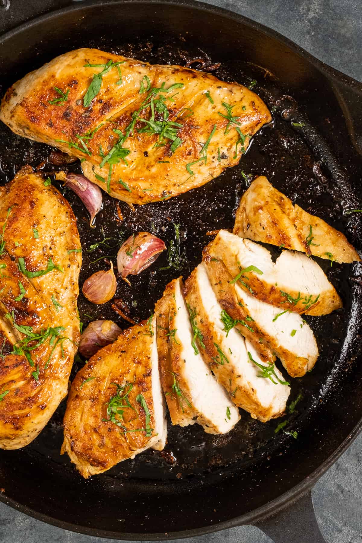 Three pieces golden brown chicken breasts, one sliced, in a cast iron pan garnished with chopped parsley and garlic cloves on the side.