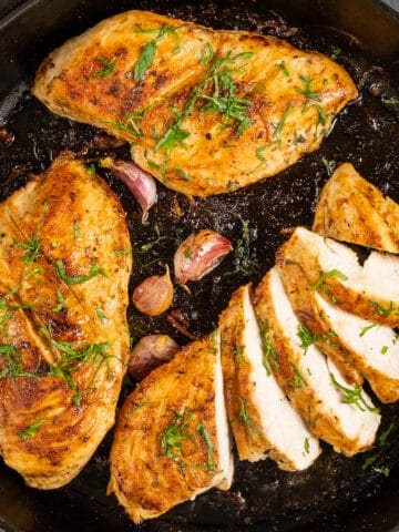 Cooked chicken breasts in a cast iron pan, one of them is sliced, garnished with parsley, accompanied by unpeeled garlic cloves.