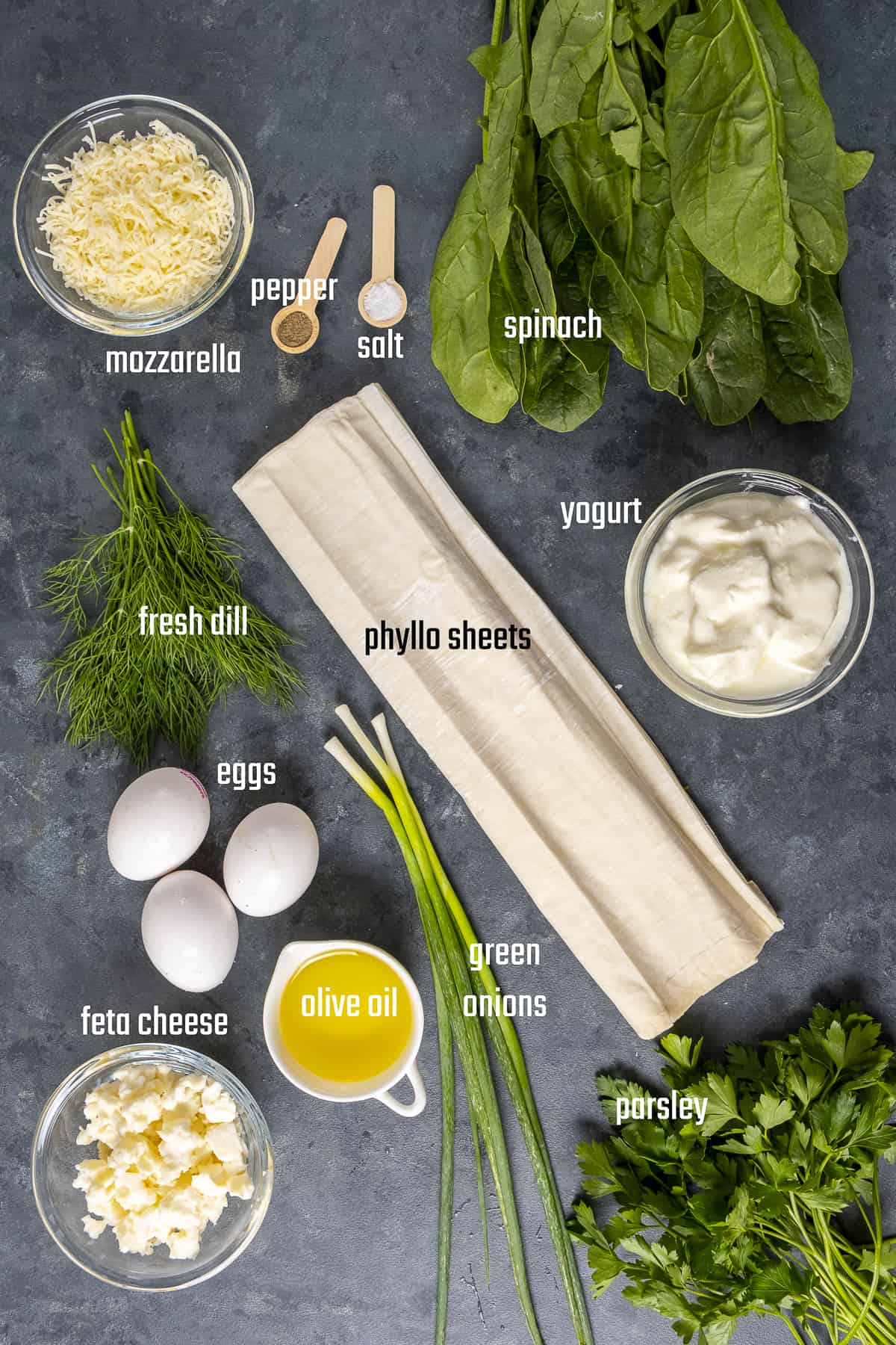 Phyllo sheets, spinach, parsley, dill, green onions, eggs, yogurt, olive oil and cheeses on a dark background.