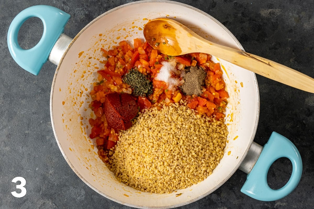 Bulgur, tomatoes, tomato paste, spices, onions and peppers in a white pan and a wooden spoon inside it.