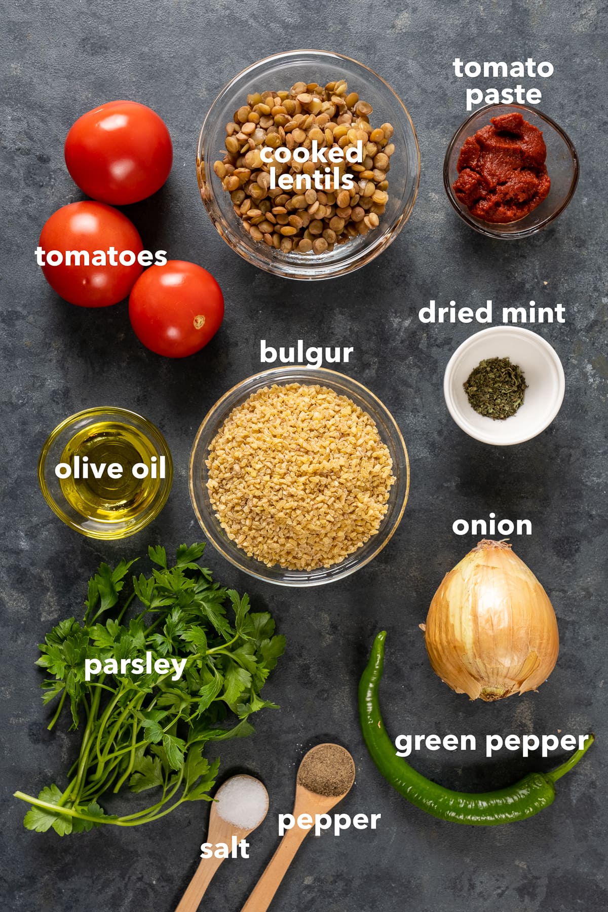 Bulgur in a glass bowl, cooked green lentils in a glass bowl, tomato paste, olive oil and dried mint in small bowls, salt and pepper in wooden spoons, onion, green pepper, parsley and tomatoes on a dark background.