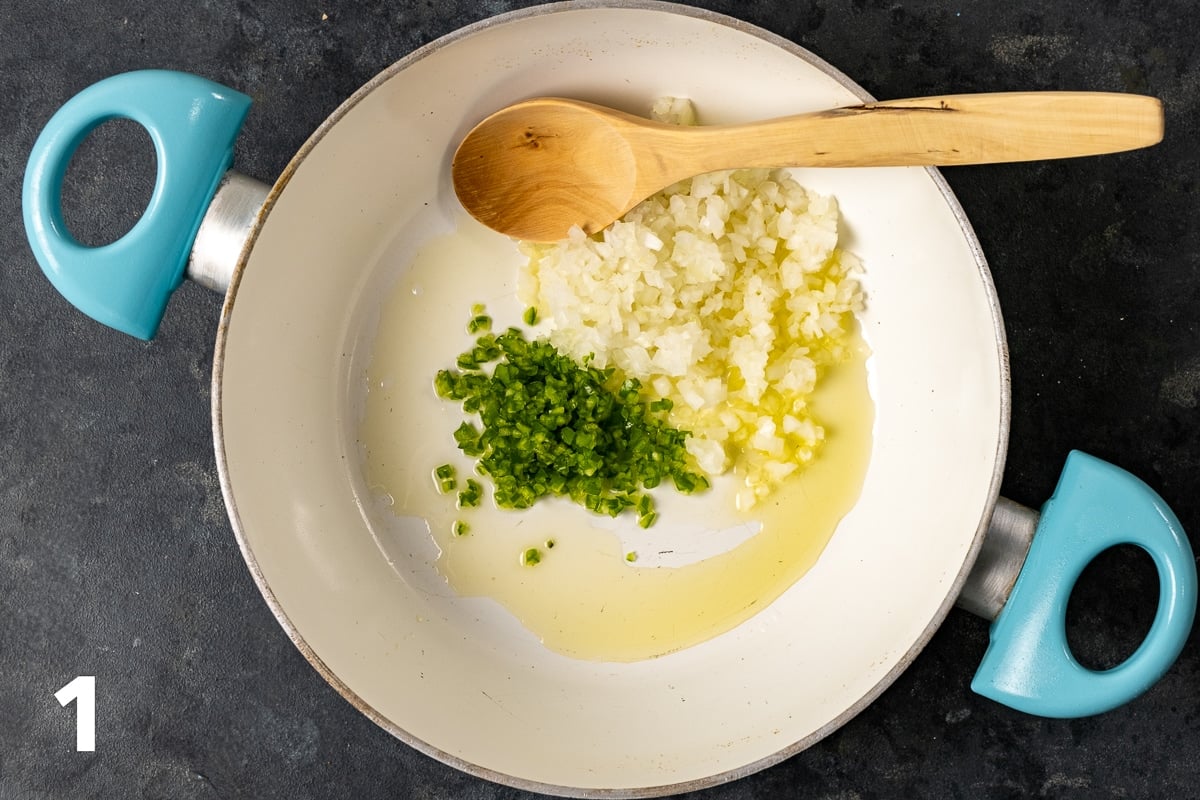 Chopped onions and green peppers with olive oil in a white pan and a wooden spoon inside it.